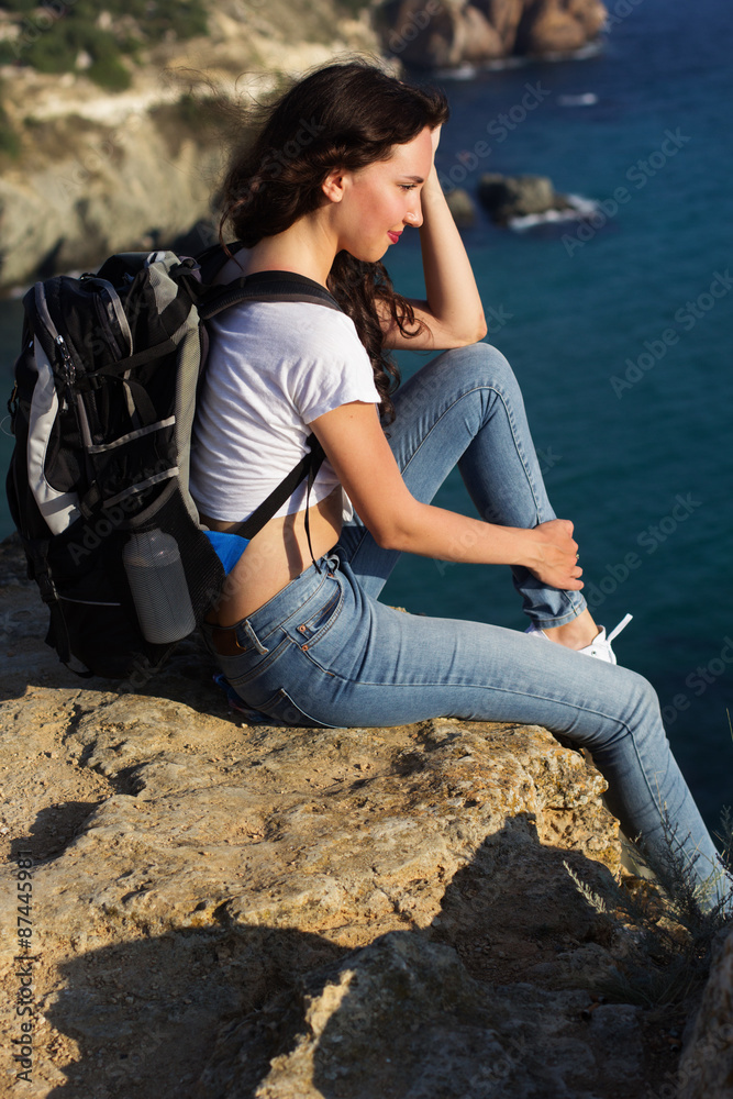 Pretty girl traveler is sitting on rock edge with backpack