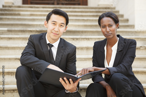 Professionally dressed man and woman sitting on an outdoor staircase make eye contact with the viewer.