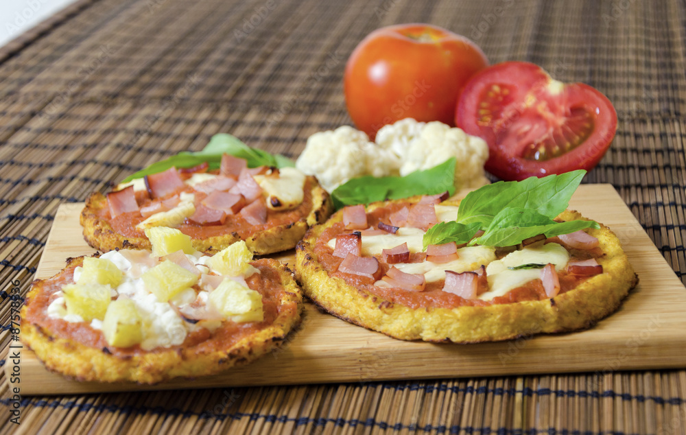 Three small round baked pizzas made of cauliflower crust and topped with bacon,ham,halloumi cheese,cottage,lountza,pineapple,turkey,tomato sauce,basil on a wooden chopping board. A healthy pizza snack