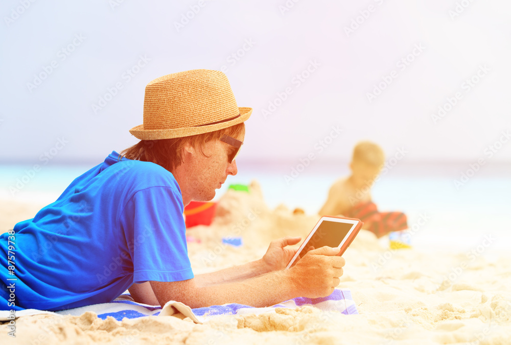 father looking at touch pad while son play on beach