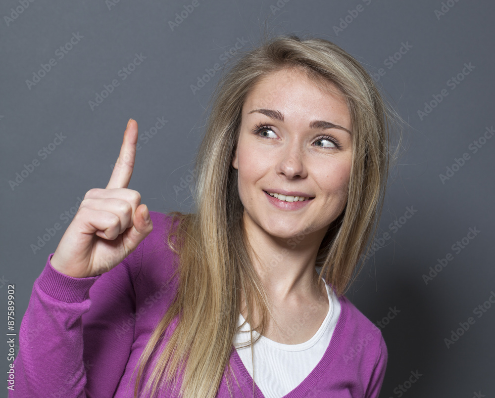 playful 20s blonde girl raising her index finger, looking at it questioning the restriction with happiness and fun - obrazy, fototapety, plakaty 