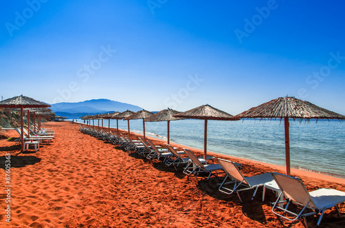 Straw umbrellas and sunbeds on a red sand beach and turquoise water.