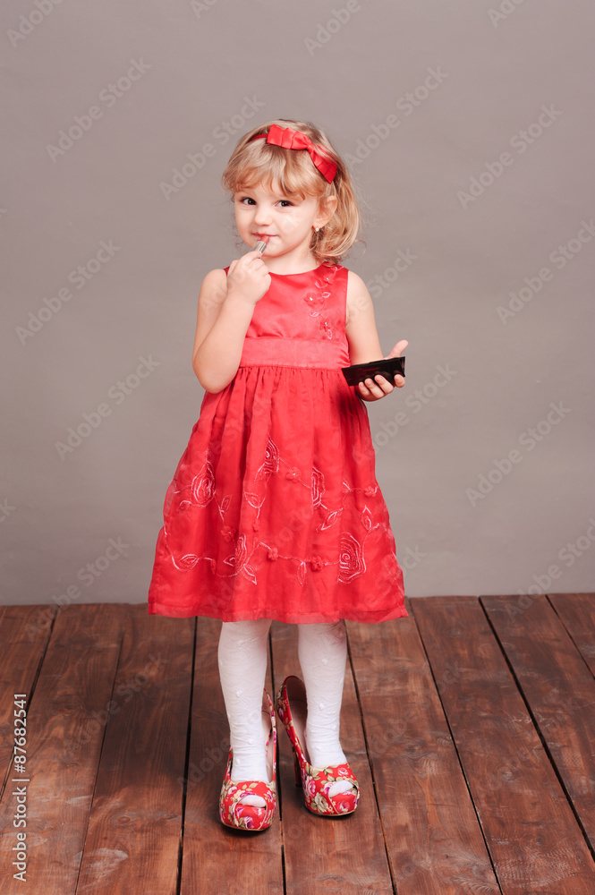 Cute little girl doing makeup, wearing mothers clothes. Portrait of little blonde girl posing in studio on gray Cute little girl doing makeup, wearing mothers clothes. Portrait of little blonde girl posing in studio on gray