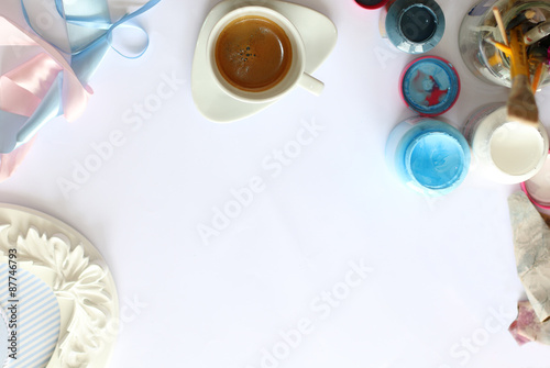 Coffee espresso in white cup, ribbons, paintbrushes, open containers of paints, sandpaper, frame on white paper  seen from above