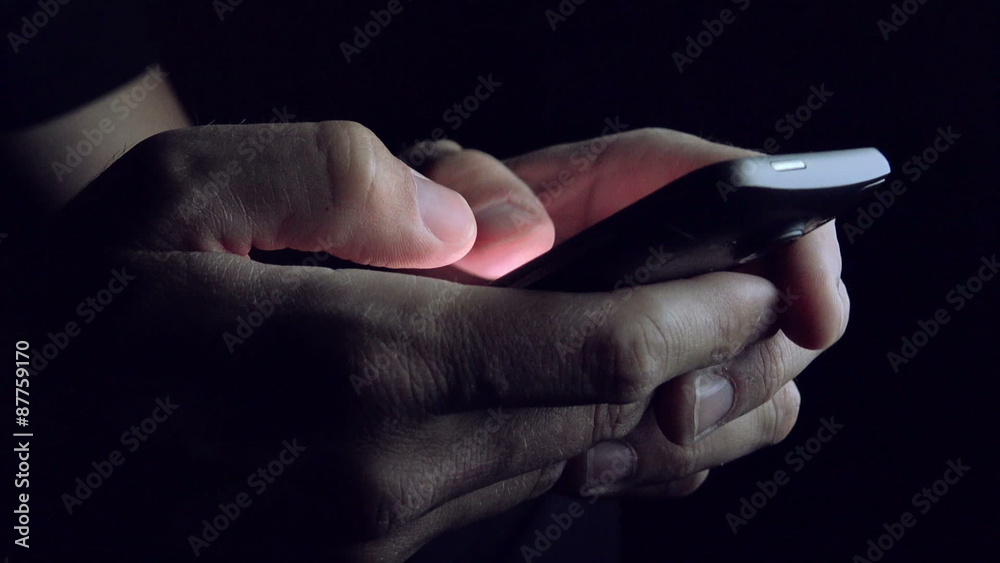 Man sending SMS text message on smartphone in dark room, close up of hands and mobile phone