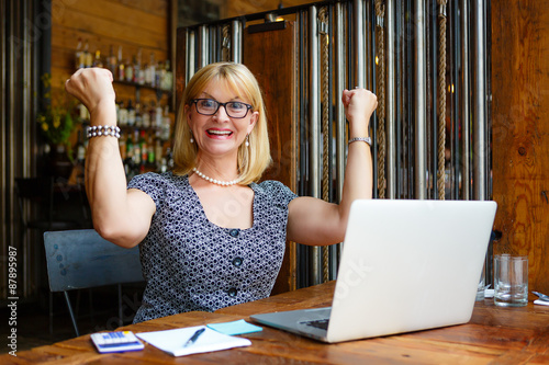 Close Up portrait Smiling old senior blonde business woman in the glasses, alone with laptop in summer cafe with gesture on face and raised hands up with fists