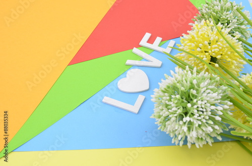 word love with love symbol in white color.vertical position with green, white and yellow artificial flower on orange, red, blue and green background