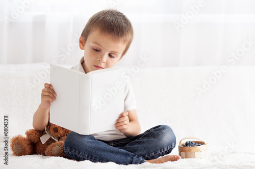 Sweet little boy, reading a book in bed and eating blueberries,
