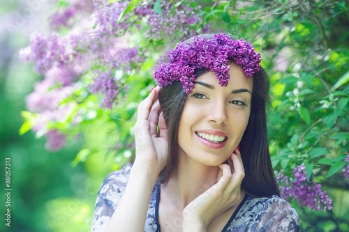 Portrait of happy and attractive young woman wearing lilac wreath posing in the blooming spring garden