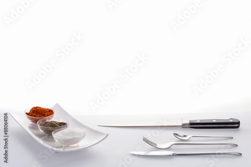 Salt, pepper, paprika in bowls in a row and a cooking knife, a knife, a fork, and a teaspoon arranged against a white background