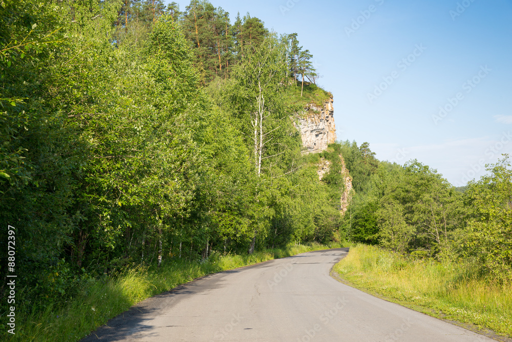 Tourist route to the Urals. Asphalt road leads to the rock in the green forest on background blue sky 