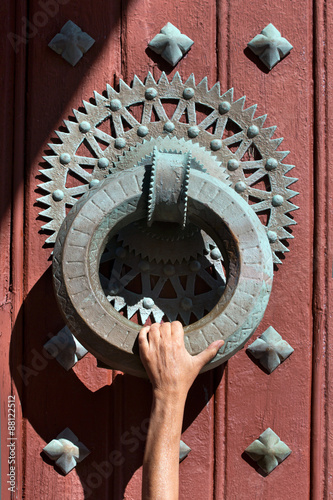 Knocker huge and heavy, very old with a hand grabs a maroon door with ornaments, all in rusty metal.