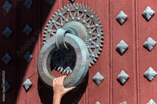 Knocker huge and heavy, very old with a hand grabs a maroon door with ornaments, all in rusty metal.