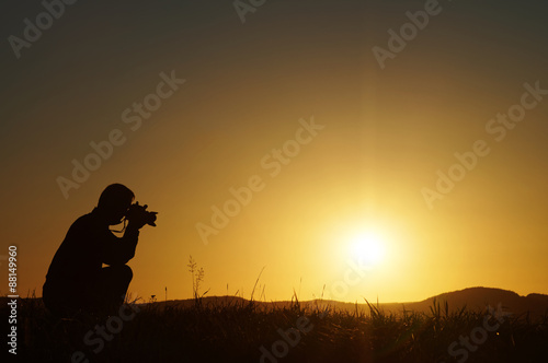 Silhouette photographer kneeling on a grassy horizon at sunset. Wooded mountains in the background.
