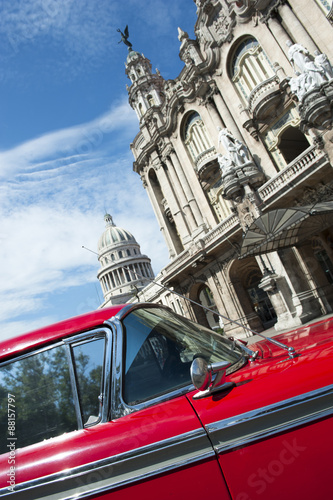 The Capitolio Building stands behind a view of the landmark architecture of the Great Theater of Havana with a bright red accent of vintage American car