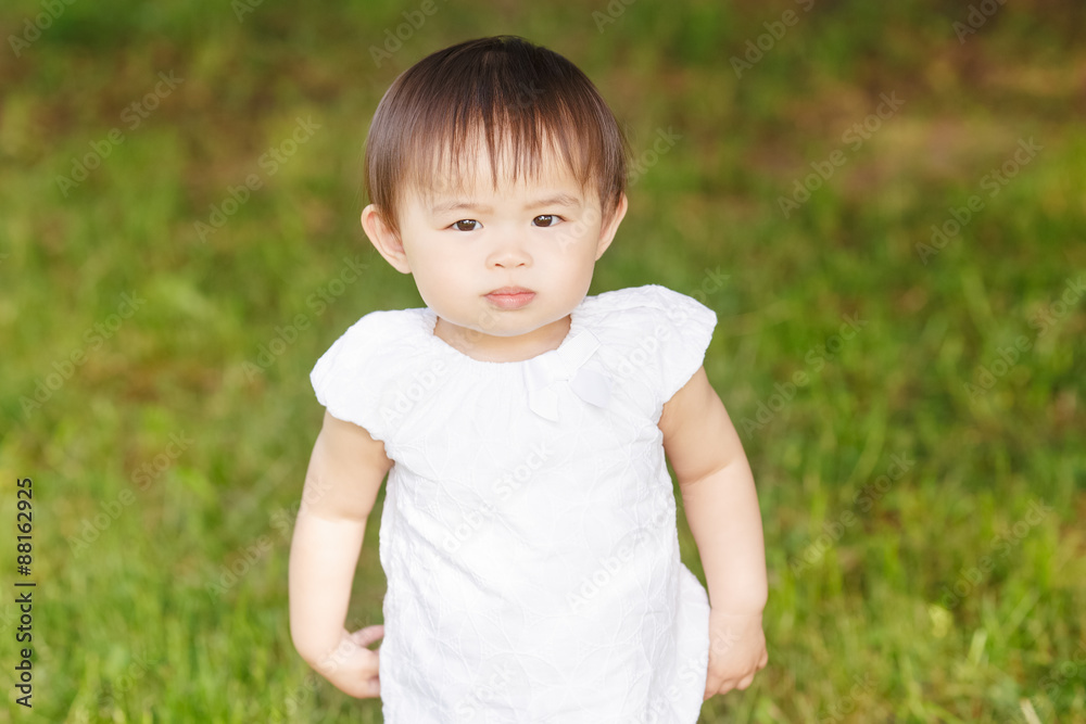 Portrait of cute adorable little Asian girl child baby one year old in white pants shirt, standing on field meadow grass on sunset playing Portrait of cute adorable little Asian girl child baby one year old in white pants shirt, standing on field meadow grass on sunset playing