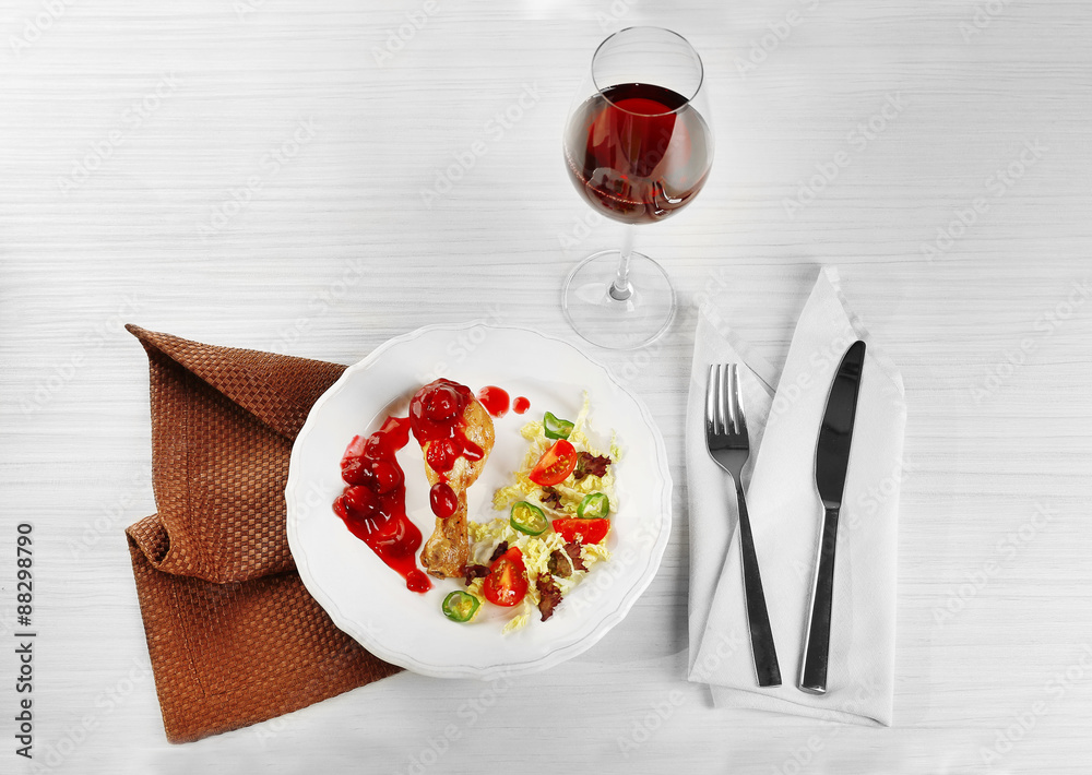 Dish of baked chicken leg and vegetable salad in white plate with glass of wine on table with napkin, closeup