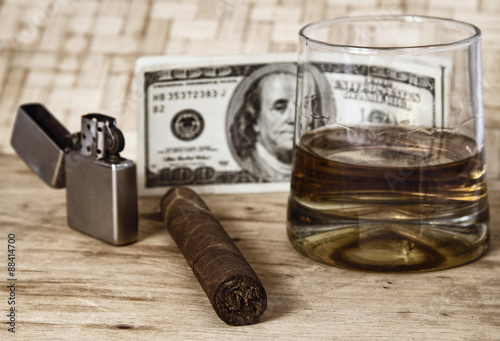 Glass of whiskey and a money with cuban cigar and lighter on a wooden table.