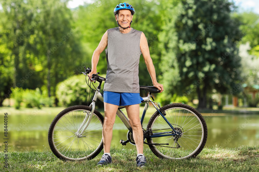 Naklejka premium Cheerful senior cyclist posing in front of his bicycle by a pond