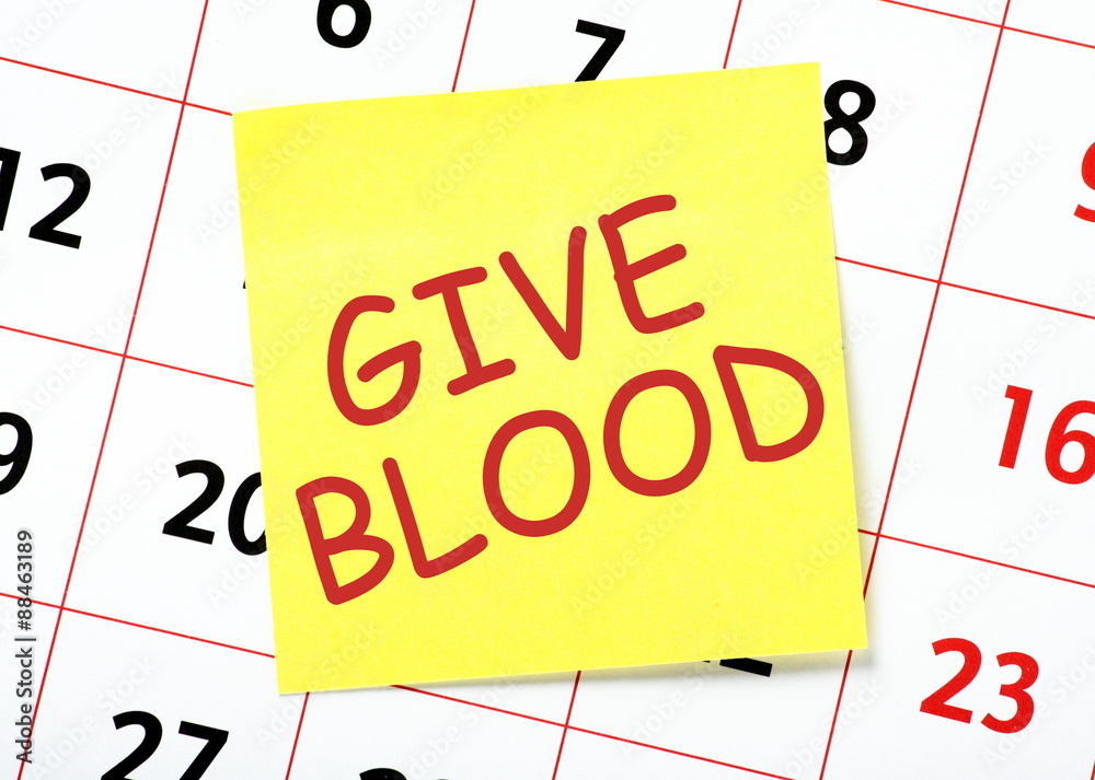 A reminder to Give Blood written in red ink on a yellow sticky note posted on a calendar A reminder to Give Blood written in red ink on a yellow sticky note posted on a calendar