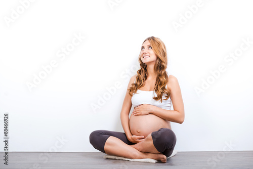 Happiness is on the way. Smiling pregnant woman touching her belly and looking up while sitting against white background