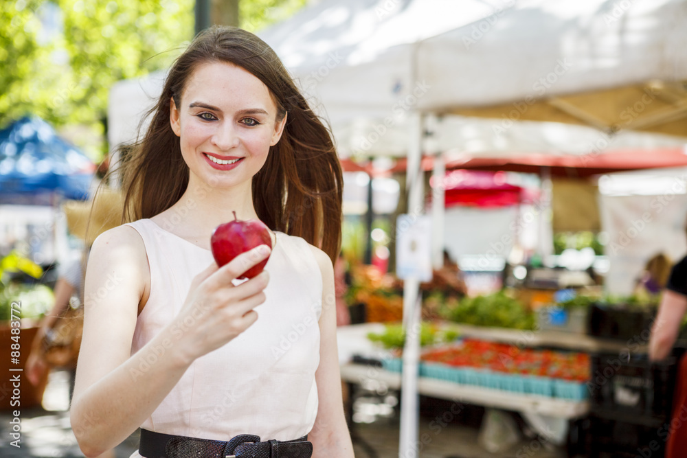 A young smiling woman holding an apple. An out of focus produce market stand is in the background. A young smiling woman holding an apple. An out of focus produce market stand is in the background.