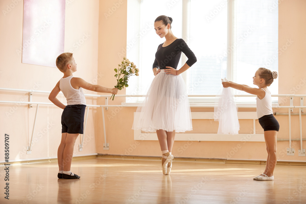 Fototapeta premium At ballet dancing class: young boy and girl giving flowers and veil to older student while she is dancing en pointe