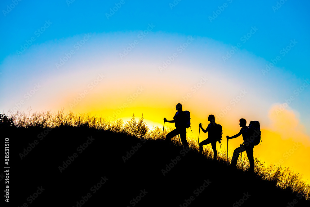 Family journey wild landscape Silhouettes of three people walking with backpacks other hiking gear up toward top of wild grass mountain mother father daughter bright luminous sunrise sky background