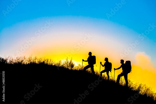 Family journey wild landscape Silhouettes of three people walking with backpacks other hiking gear up toward top of wild grass mountain mother father daughter bright luminous sunrise sky background