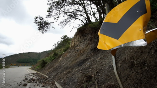 Panning shot of a broken and crumbled curve in the road warning sign in front of rocks strewn all over the road from what appears to be the scene of a motor vehicle accident.