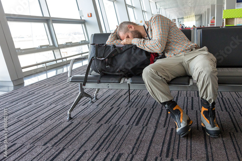 Man stuck at airport.
Image of bearded hippy style dressed person sleeping on his travel backpack inside airport waiting lounge sitting in black chair with heavy alpine boots on his legs