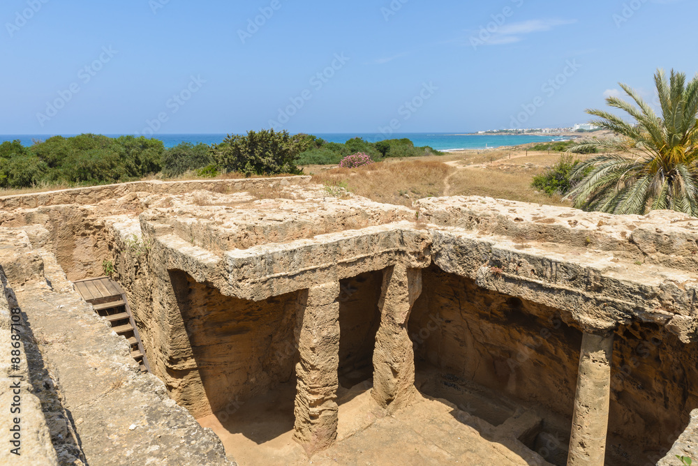 Looking down into a chamber of an ancient carved stone burial tomb at an archaeological site in Paphos, Cyprus with the blue Mediterranean Sea in the background.