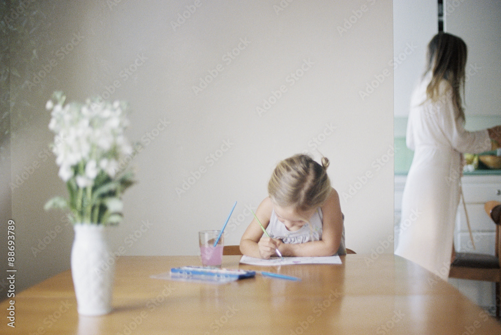 Young girl sitting at a table, painting, a vase with white flowers, a woman in the background. Young girl sitting at a table, painting, a vase with white flowers, a woman in the background.