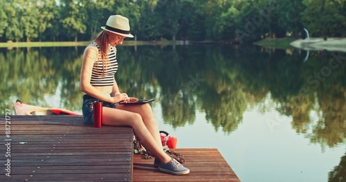 Woman using digital tablet sitting on a wooden jetty by the lake, back view. Slow Motion, 4K. Happy young girl browsing Internet, using app and communicating outdoors. Relax and technology.