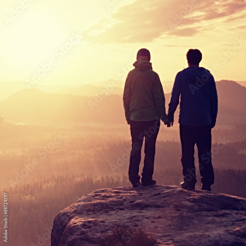 Couple in love enjoying tender moments during sunset . Young pair of hikers hand in hand on the peak of rock empires park and watch over the misty and foggy morning valley to Sun.