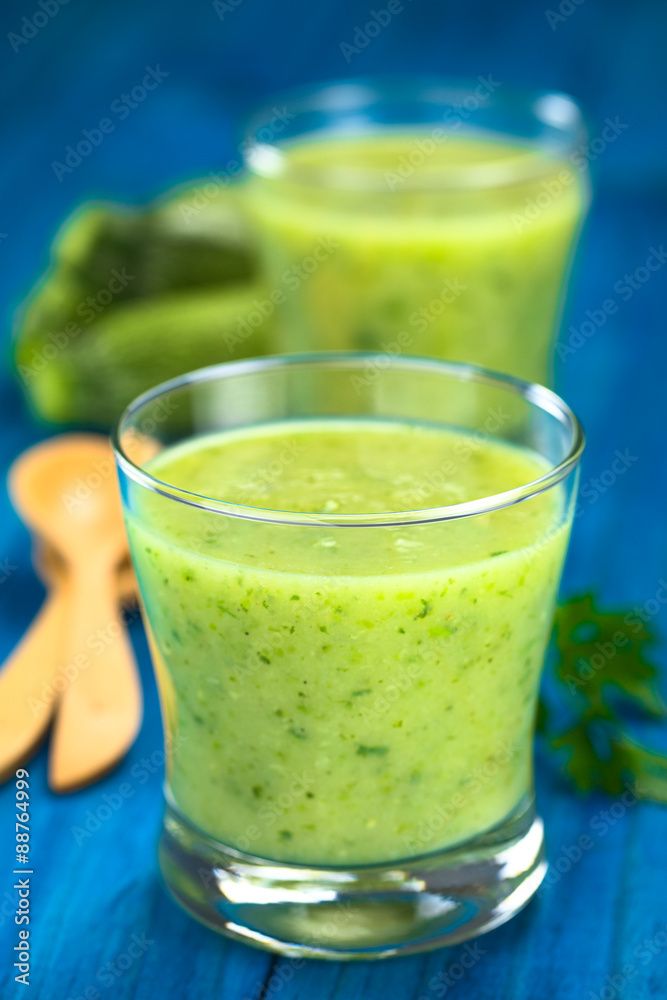 Zucchini cream soup served in glasses on blue wood with wooden spoon, parsley and small zucchini in the back (Selective Focus, Focus on the front of the rim of the first glass) Zucchini cream soup served in glasses on blue wood with wooden spoon, parsley and small zucchini in the back (Selective Focus, Focus on the front of the rim of the first glass)