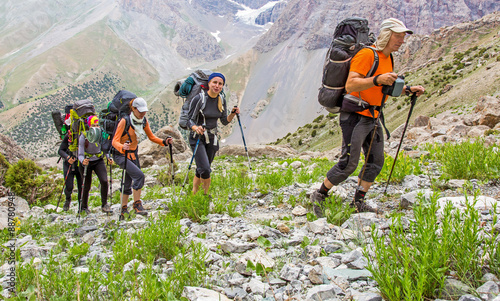 People traveling in mountains.
Large group of tourists of different sex ethnic nation race age young and old man woman walking up on rocky path with green grass forest and mountain peaks around