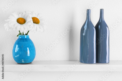 Decorative vases with flowers on wooden shelf on white wallpaper background