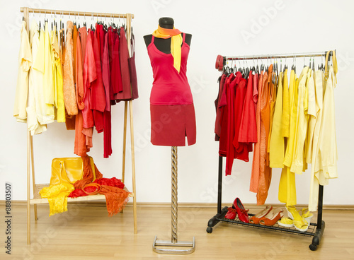 Wardrobe with yellow, orange and red clothes arranged on hangers and a red outfit on a mannequin. Dressing closet with bright color coordinated clothes on hangers, shoes and accessories.