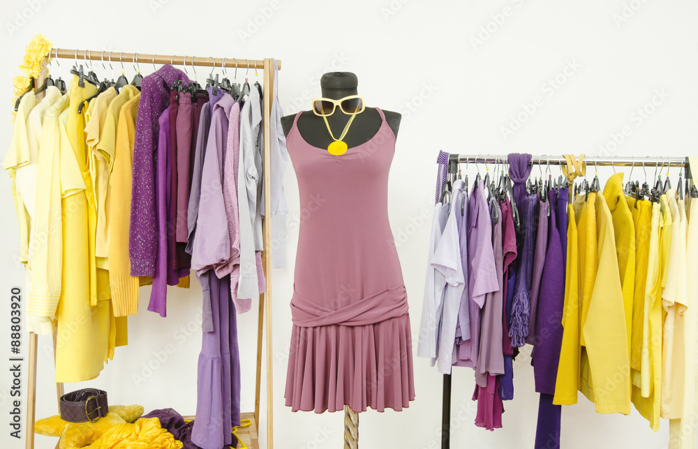 Dressing closet with complementary colors violet and yellow clothes. Wardrobe with purple and yellow clothes arranged on hangers and a summer dress on a mannequin.