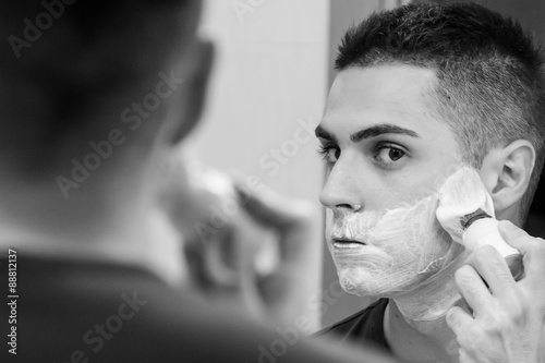 Handsome young man is shaving his face with shaving brush and looking at the mirror