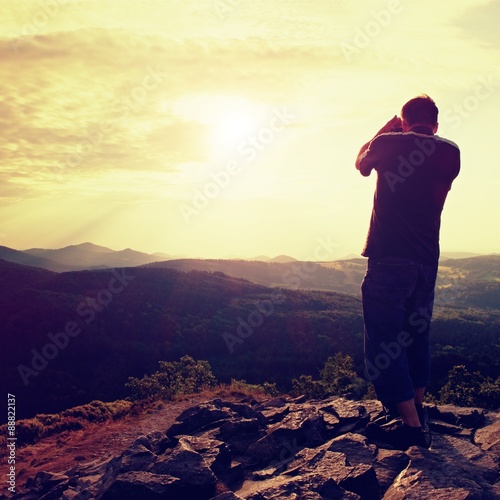Professional photographer in jeans and shirt  takes photos with mirror camera on peak of rock. Dreamy landscape, orange  Sun at horizon