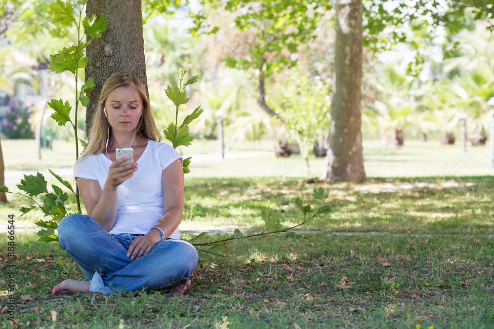 A beautiful blonde woman sitting in a park and surfing in the net with her smartphone A beautiful blonde woman sitting in a park and surfing in the net with her smartphone
