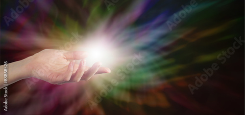 Fototapeta Naklejka Na Ścianę i Meble -  Female Healer with hand out palm up with a ball of white energy appearing to manifest on a dark rainbow colored energy formation background banner with plenty of copy space