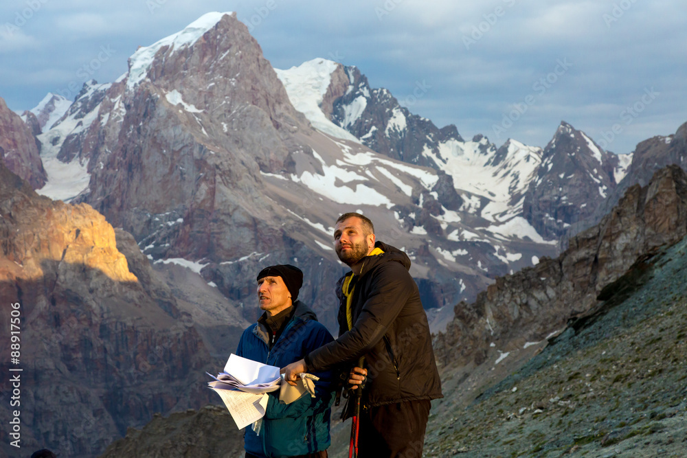 Climbers learning future route of ascent.
Two people old and young man discuss mountain way keeping stack of papers with route description guidebook printed faces highlighted last sunbeams warm sun