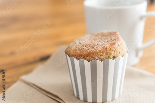 A fresh, sugar topped muffin with a striped wrapper and a white cup of coffee on a rustic wooden cafe table.