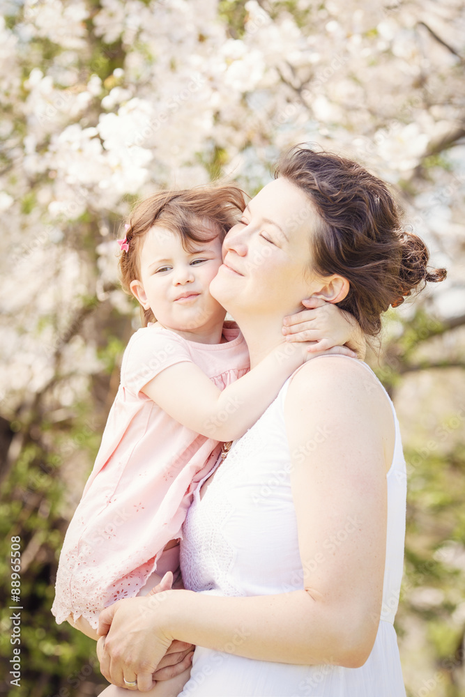 Portrait of caucasian pregnant mother in long white dress holding and kissing her daughter in pink clothes on spring summer day in park outside among blooming cherry trees Portrait of caucasian pregnant mother in long white dress holding and kissing her daughter in pink clothes on spring summer day in park outside among blooming cherry trees