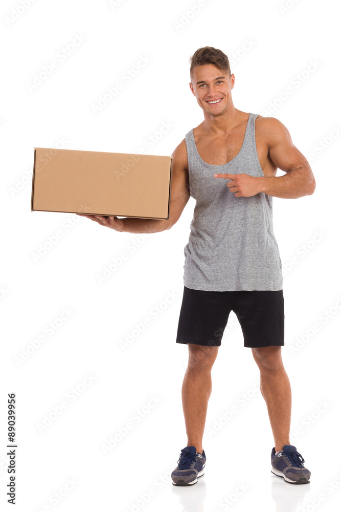 This Is Your Delivery. Smiling young man in sorts clothes standing with legs apart, holding carton box in one hand and pointing. Full length studio shot isolated on white. This Is Your Delivery. Smiling young man in sorts clothes standing with legs apart, holding carton box in one hand and pointing. Full length studio shot isolated on white.
