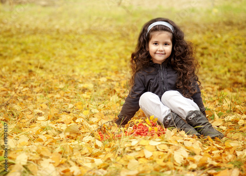 little cute girl with dark curly hair sitting in the autumn forest with a bouquet of leaves