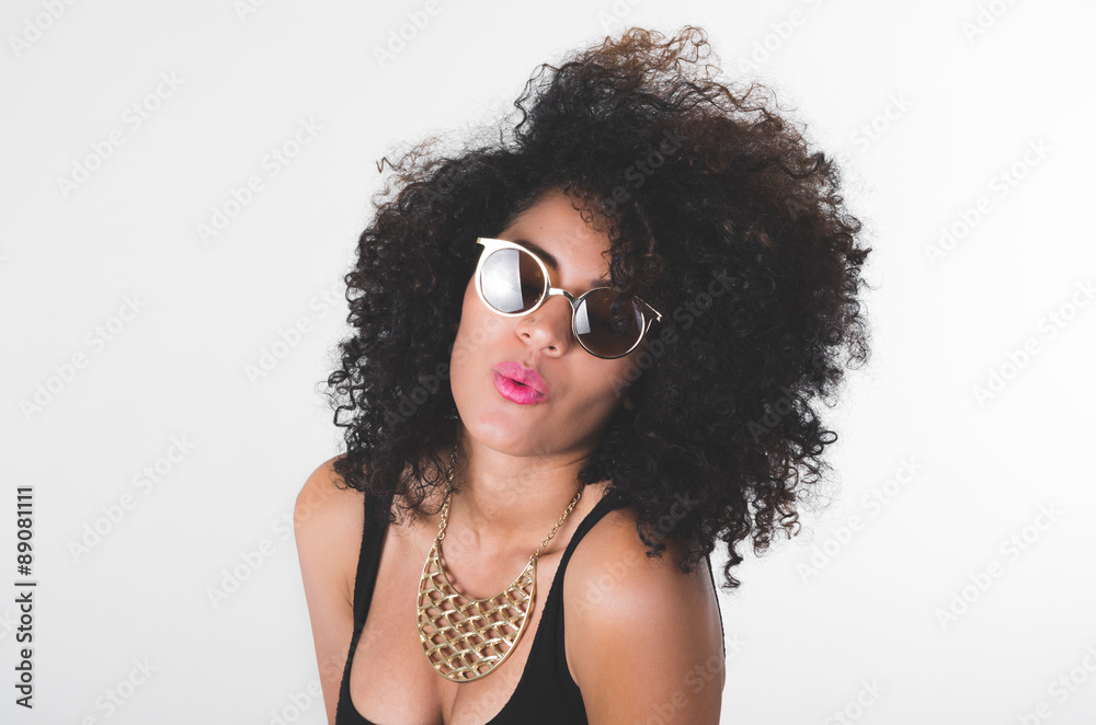 Fototapeta premium Headshot hispanic model wearing black dress, golden necklace, sunglasses and curly hair making kiss lips for camera